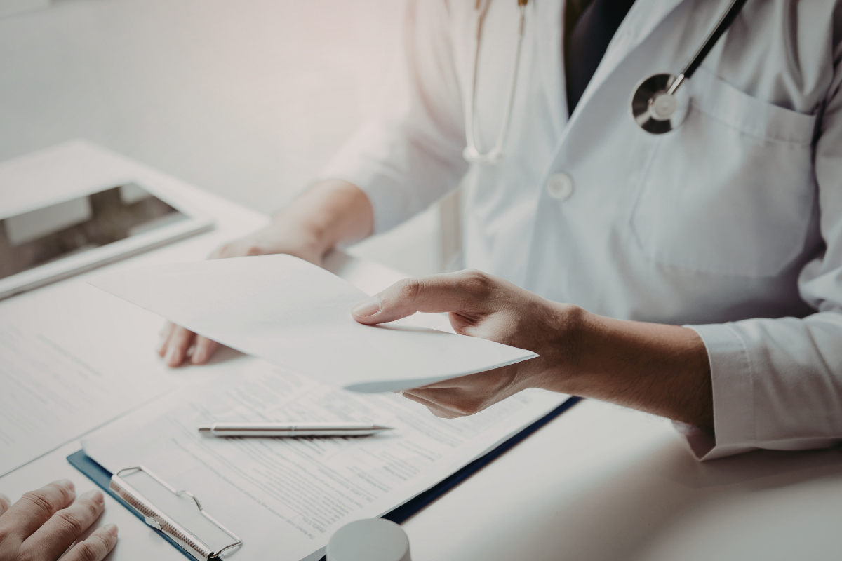 Doctor in a white coat with a stethoscope hands a patient documents over a desk with a pen and clipboard nearby.