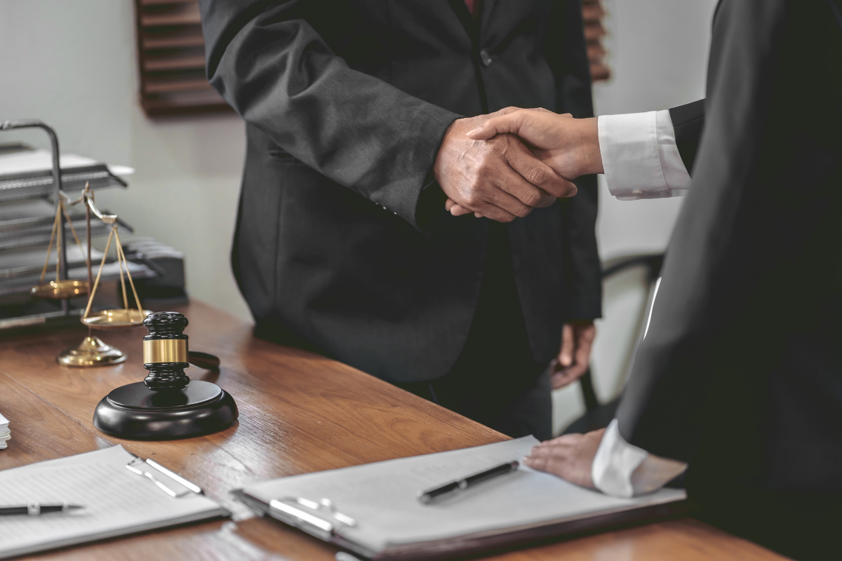 Two professionals in suits shake hands over a wooden desk in a law office, with a gavel and legal papers nearby.