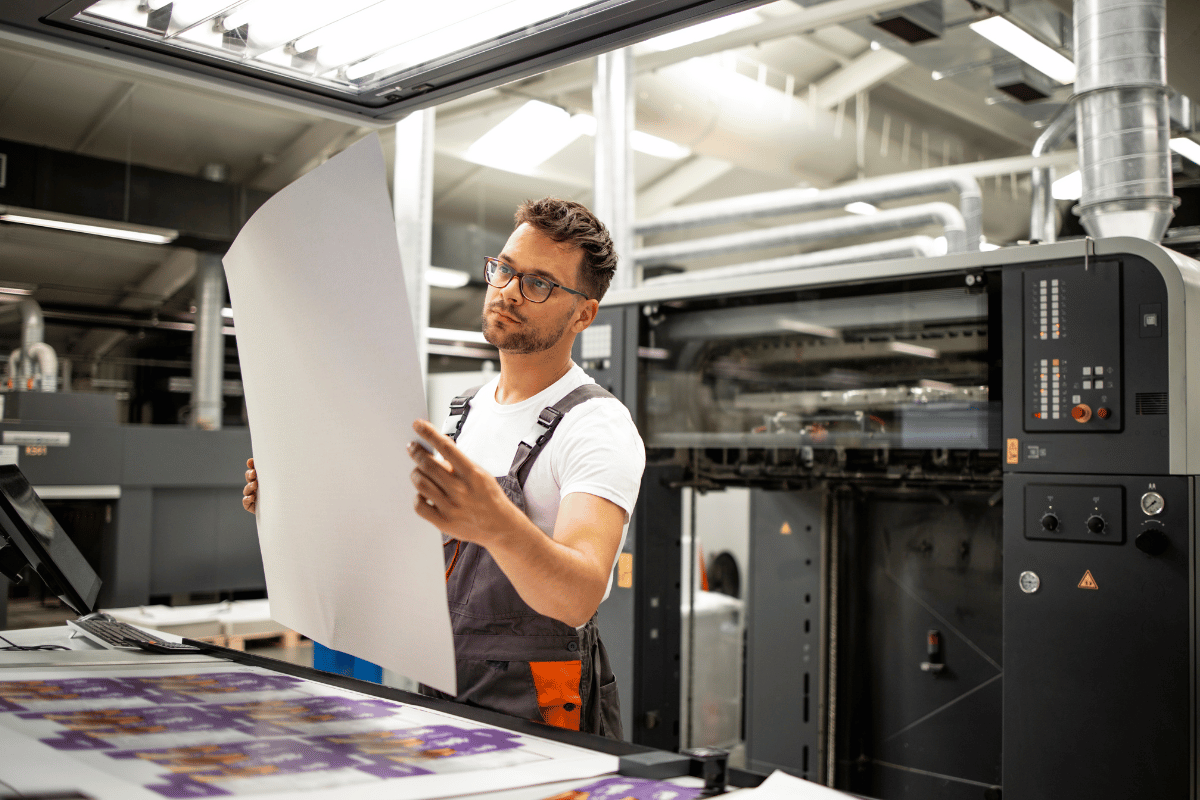 Maintenance worker inspects a large sheet of paper beside an industrial printer in a printing facility.